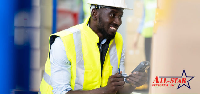 An African American male is wearing a yellow safety vest, a hard hat, and is holding a warehouse scanner in his hands.