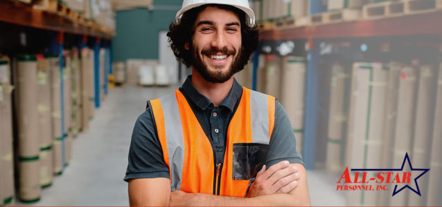 A white male is wearing an orange safety vest in a warehouse setting. He is also wearing a white hard hat, has his arms crossed and is smiling.