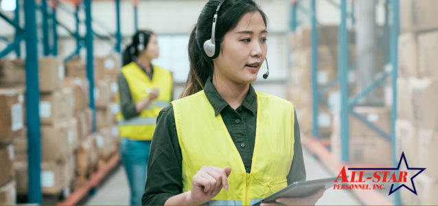 A female of Asian decent is talking into a headpiece and carrying a clipboard or tablet. She is wearing a dark green button down shirt and a yellow safety vest.